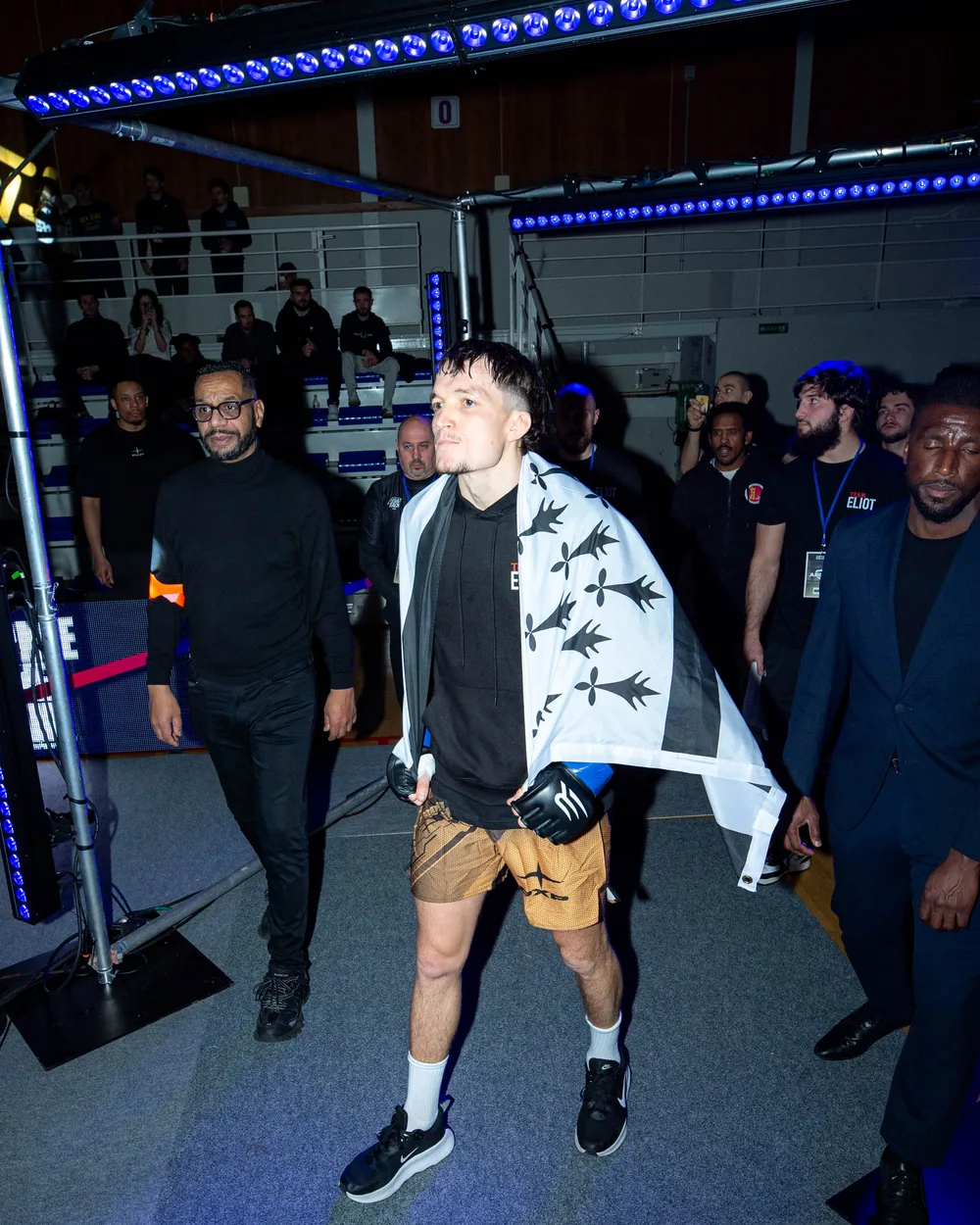 Entrée d’un combattant drapé dans un drapeau breton, encadré par son staff, sous les néons bleus de la salle