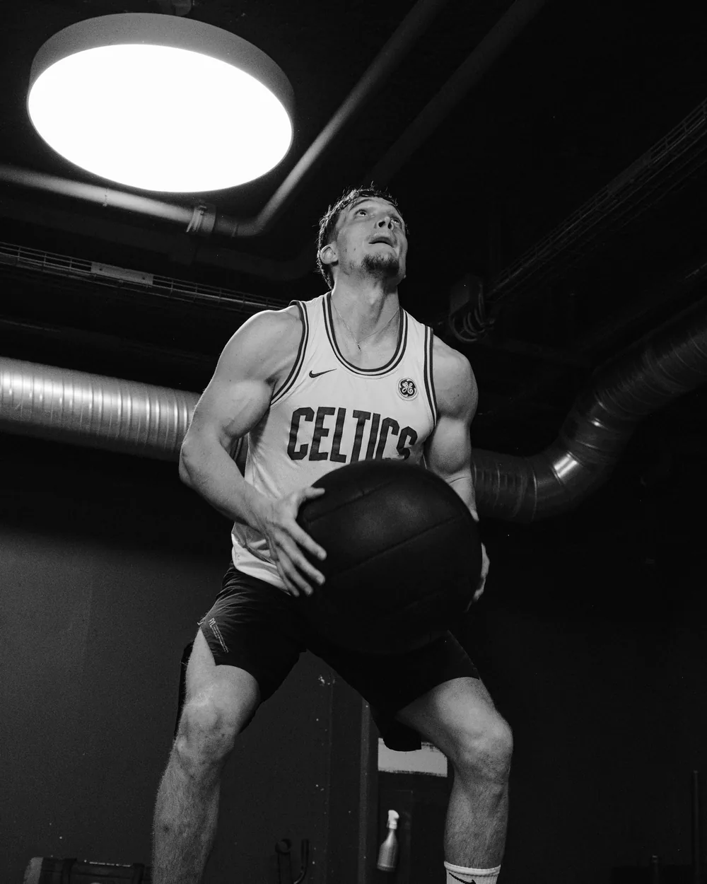 Basketteur en maillot, ballon calé contre lui, sous un plafond de parking, en noir et blanc
