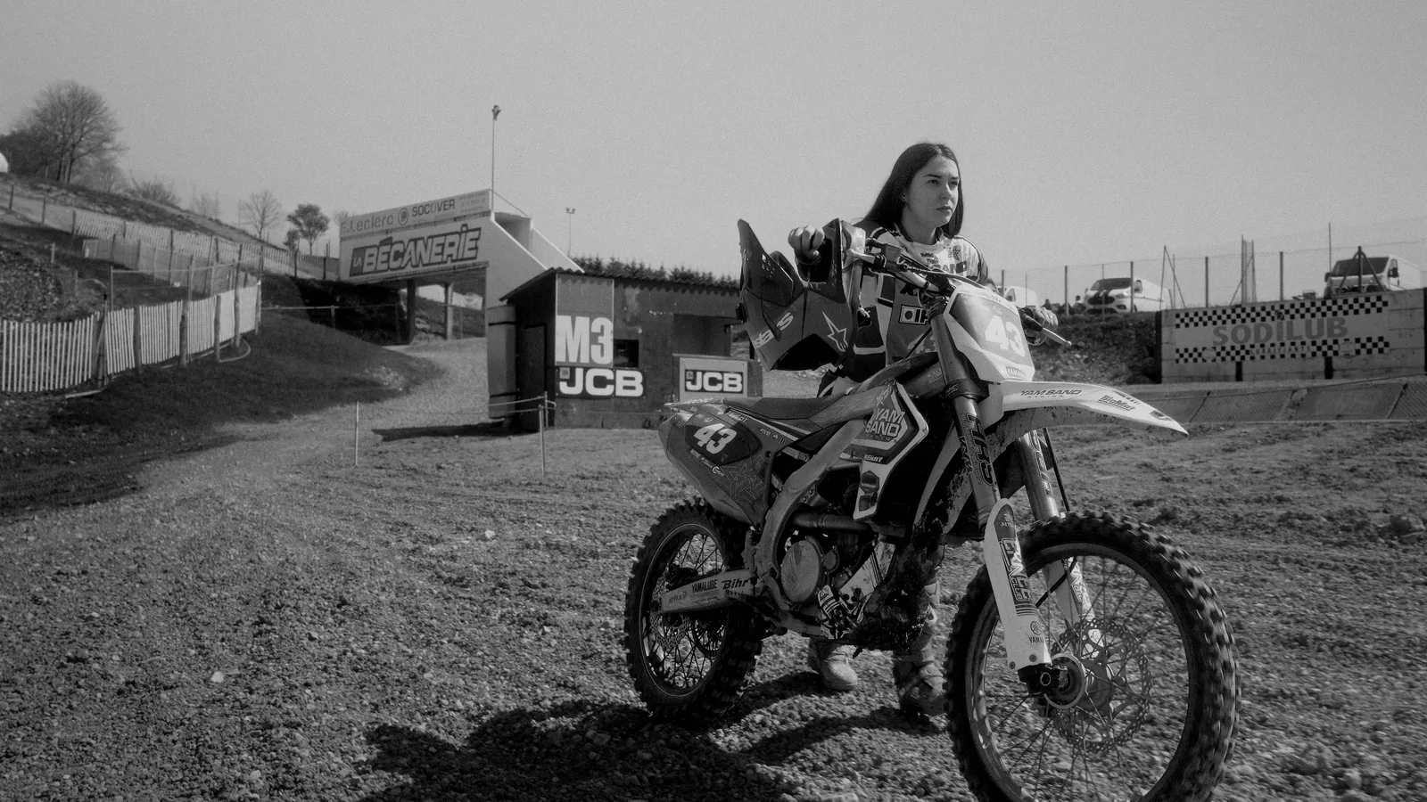 Pilote de motocross à l’arrêt sur la piste, panneaux de circuit en arrière-plan, en noir et blanc